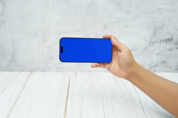 Hand holding smartphone with solid blue screen in landscape orientation on bright wooden desk and light wall background. Concept of mobile mockup, app demo, video advertising, and digital branding.