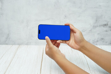 Hand holding smartphone with solid blue screen in landscape orientation on bright wooden desk and light wall background. Concept of mobile mockup, app demo, video advertising, and digital branding.
