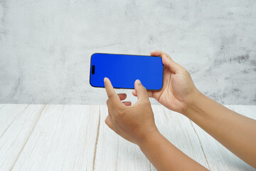 Hand holding smartphone with solid blue screen in landscape orientation on bright wooden desk and light wall background. Concept of mobile mockup, app demo, video advertising, and digital branding.
