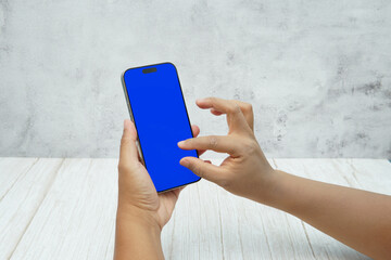 Hand holding smartphone with solid blue screen on bright wooden desk and light wall background. Concept of mobile mockup, app UI presentation, digital marketing, and modern technology.