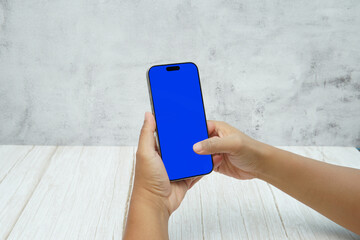 Hand holding smartphone with solid blue screen on bright wooden desk and light wall background. Concept of mobile mockup, app UI presentation, digital marketing, and modern technology.