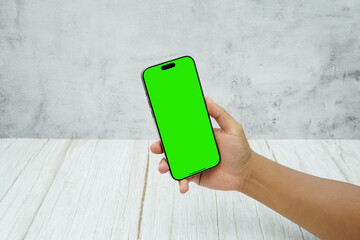 Hand holding smartphone with green screen on bright wooden desk and light wall background. Concept of mobile mockup, app presentation, digital marketing, UI UX design, and screen replacement.