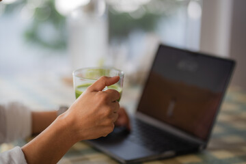 Close-up image of a male holding a glass of iced matcha green tea, sipping iced green tea and working on laptop while relaxing at a cafe.