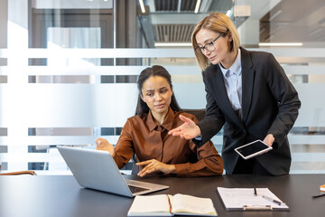 Two professional business women collaborating on a laptop project during a corporate meeting, discussing strategy and reviewing digital documents in a modern office workplace