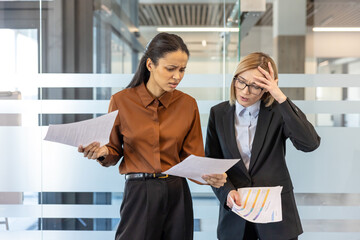 Two diverse businesswomen reviewing documents with concerned and frustrated expressions, indicating financial stress, complex problems, or unexpected challenges in their corporate office environment