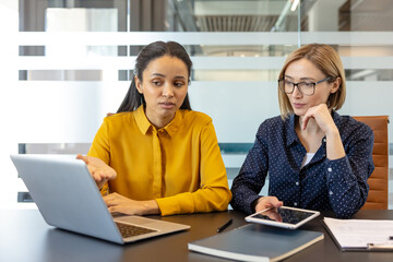Two diverse businesswomen discussing data on a laptop in a modern office, one presenting information while the other listens intently, showcasing collaboration and mentorship
