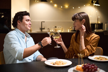 Caucasian young man and Caucasian young woman sitting at table clinking wine glasses during dinner, smiling and making eye contact, plates of food and fruit on table