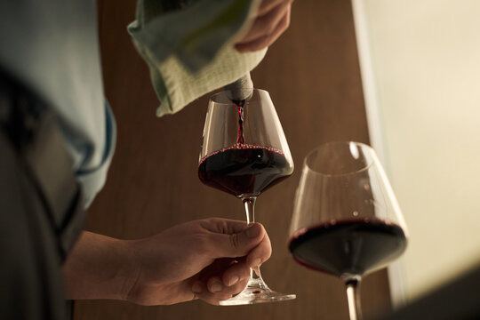 Caucasian middle aged man pouring red wine into glass, holding stemware with one hand while using towel to steady bottle, close up of hands and wine glasses in indoor setting