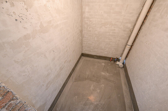 Unfinished bathroom corner with plastered walls, concrete floor, exposed plumbing pipe, water supply valves, hydroisolation layer visible, awaiting tiling and fixtures.