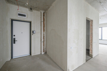 Unfinished apartment interior with plastered walls, concrete floor, exposed brick sections, white entrance door, visible plumbing pipes, open doorway to adjacent room, minimal fittings.