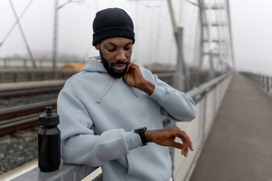 Thoughtful Black male runner checking his pulse on a smartwatch on an urban bridge, wearing a hoodie and beanie, water bottle in hand, foggy morning workout focused on recovery and motivation