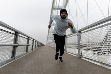Obraz premium Low-angle view of a Black male runner sprinting on a foggy urban bridge, holding a water bottle. Dynamic motion, cardio training, endurance and focus in modern outdoor fitness scene