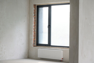 Fototapeta premium Bare room with unfinished walls; large black-framed window with brickwork visible on one side; white radiator mounted below window; minimalistic, empty space, natural light.