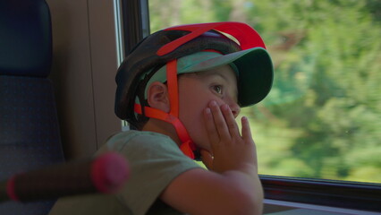 Little boy wearing bicycle helmet and cap rests his face on his hand while gazing pensively out train window, seated indoors with blurred summer landscape outside