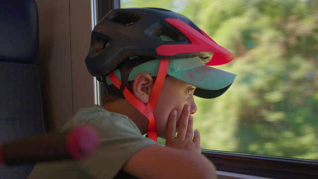 Side profile of young boy in bike helmet leaning on train window with fingers on face, observing green forest pass by outside while traveling in daytime