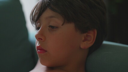 Young boy with serious expression resting on armchair indoors, natural light from window highlighting quiet mood and focused gaze © Marco