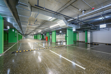 Fototapeta premium Modern underground parking with polished concrete floor, green pillars, exposed ducts, fluorescent lighting, marked lanes, speed bumps, numbered spaces (e.g., 1024), clean and well-lit.