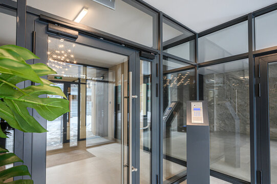 Modern entrance with glass doors, access control panel, sleek metal framing, indoor plants, polished floor, stylish lighting, clean minimalist design.
