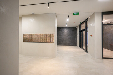 Obraz premium Modern residential lobby with light-toned tiled floor, beige walls, dark brick accent wall, wooden mailboxes, glass doors, track lighting, and exit sign — sleek and functional