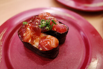 An overhead close-up shot of two pieces of gunkan maki sushi filled with marinated spicy tuna and topped with chopped green onions, served on a speckled red ceramic plate. © Peeradontax