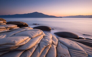 Sunset over calm sea, layered rocks in foreground, distant mountains under soft gradient sky.