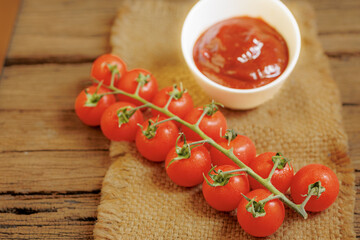 Fresh cherry tomatoes on vine rest on rustic wooden table beside small bowl of vibrant red tomato sauce, creating warm and inviting culinary scene