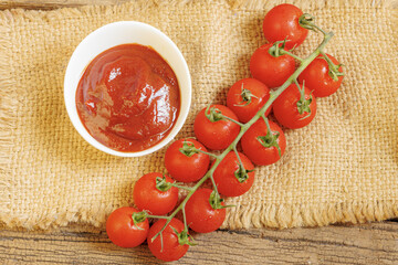 Fresh cherry tomatoes on vine next to bowl of rich tomato sauce on rustic burlap surface, creating natural and appetizing presentation