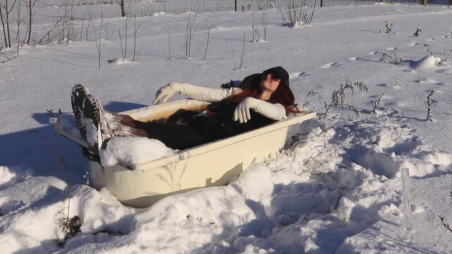 Young woman in old bathtub in the middle of a snowy field enjoying the winter sun, humorous atmosphere, freedom, relaxation and creative winter mood.