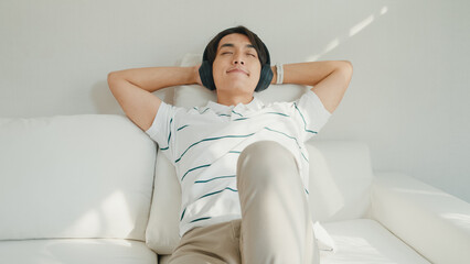 Youth attractive Asia male with casual cloth calm feeling lay down on sofa close eyes wear headphone listen music playlist relaxing at living room in cozy house. Lifestyle leisure at home concept.