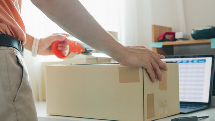 Close up young Asia male entrepreneur E-commerce small business prepare product put in paper box hands hold packing machine and sealing cardboard boxes with duct tape at small online retail shop.