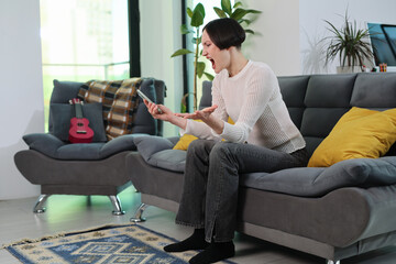 Obraz premium A young woman sits on a stylish couch, visibly upset as she engages with her smartphone. Her expressive face shows frustration in a bright, modern living room filled with plants and soft pillows.