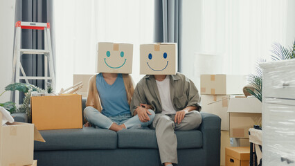 Happy Asian couple sit on sofa couch used carton box made smiling face funny dance front of camera in living room at new house. Husband and wife moving new house, Apartment Rent And Ownership Concept.