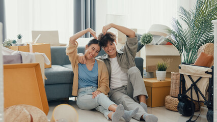 Happy Asian couple sit on floor in front of sofa couch smile looking at camera making heart sign in living room at new house. Husband and wife moving new house, Apartment Rent And Ownership Concept.