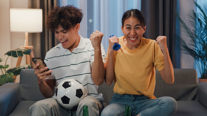 Asian new marriage couple sit on sofa excited cheering watching football sport game on television in living room at home. Young couple sport fans hold football watching online score on smartphone.