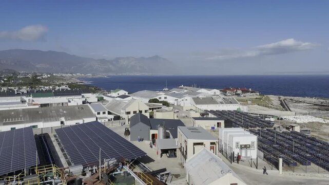 Drone flies east between abalone farm and solar panels to the new harbor on sunny day in Hermanus on the Garden Route in South Africa