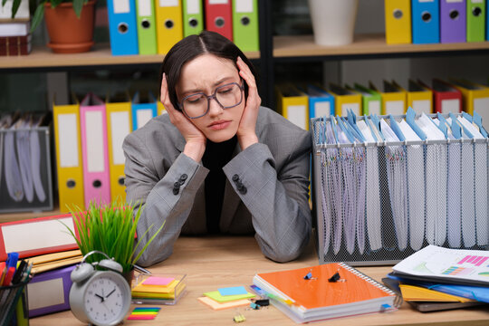 A stressed office worker sits at a cluttered desk with piles of documents and colorful sticky notes. With hands on her head, she battles the pressure of tight deadlines in her vibrant workspace.