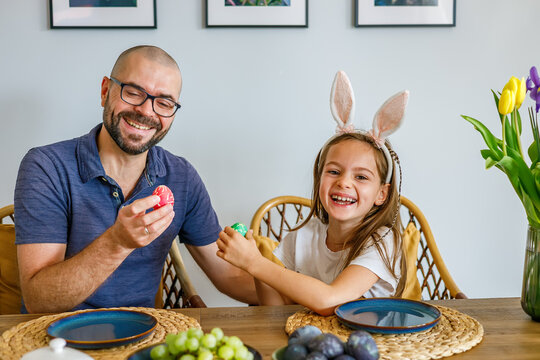 Happy father and young daughter with bunny ears laughing while playing traditional easter egg tapping game at wooden dining table in bright modern home interior
