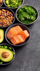 Top-down view of small bowls with salmon, avocado, greens, nuts, and seeds on a dark stone surface, glossy hue