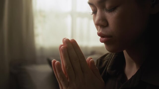 Close-up view of serene young woman with closed eyes and folded hands praying in morning with soft light filtering through window in background