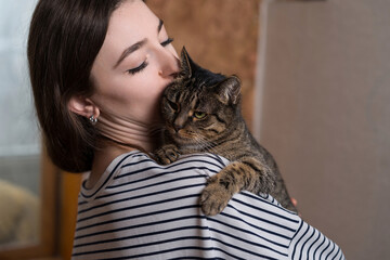 Close-up of a young caucasian woman hugging and kissing a cute, friendly, fluffy domestic cat. Pet,...