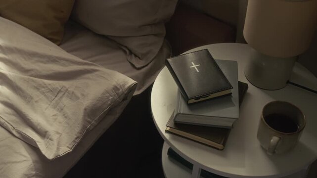 Books, leather bible, tea mug and lamp on white round bedside table next to unmade bed with grey blanket and two pillows