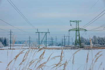 Snow-covered landscape featuring high-voltage power lines and transmission towers against a cloudy sky with tall grass in the foreground