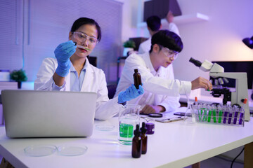 Asian female scientist holding a test tube with a solution in gloves in a research lab or doing chemical experiments, Group of young scientists in lab coats collaborate in a high-tech laboratory