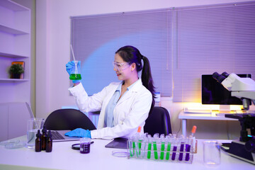 woman scientist wearing lab coat and gloves adding reagent to beaker for chemical experiment using dropper while sitting at desk with microscope and test tubes in laboratory