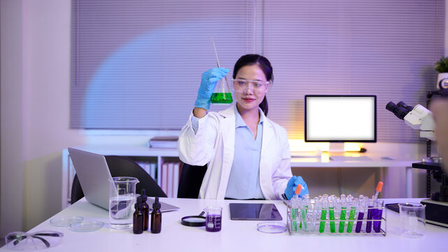 woman scientist wearing lab coat and gloves adding reagent to beaker for chemical experiment using dropper while sitting at desk with microscope and test tubes in laboratory - Powered by Adobe