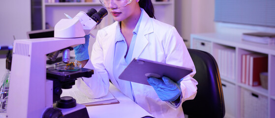 A female scientist using a microscope for research in a modern lab. She is focused and dressed in a lab coat holding tablet, gloves, and safety glasses