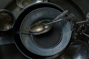 Moody close-up of antique pewter kitchen utensils: stacked metal sieves, vintage silver spoons, sugar tongs, and perforated strainer arranged on a dark textured surface. 