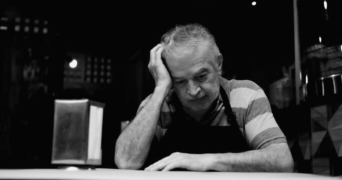 Older small business owner resting head on hand behind caf&eacute; counter conveying worry financial strain fatigue and quiet anxiety inside struggling neighborhood caf&eacute; in black and white