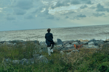 Figure in dark jacket on a rocky shore gazing at the sea, tranquil horizon and cloudy sky, coastal...