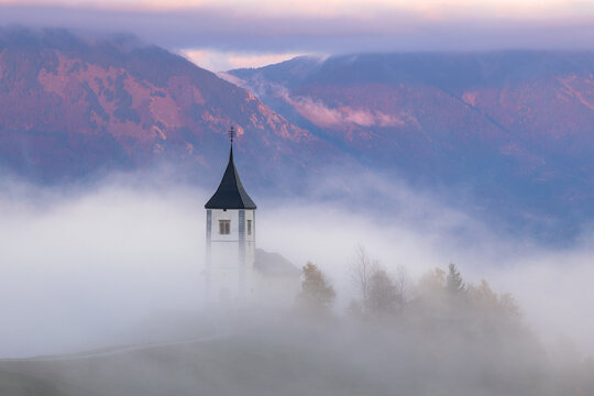 Aerial view of a church spire piercing through a sea of ethereal fog, nestled against the backdrop of rugged mountains, Spodnja Lua, Slovenia.